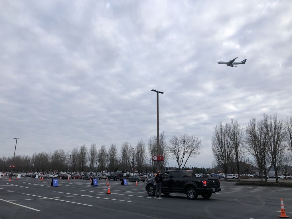 Plane visible overhead as vehicles que in airport parking lot.