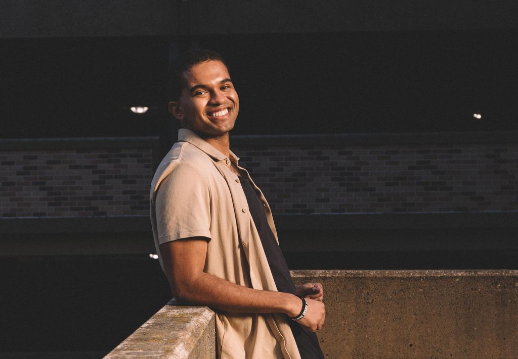 A young man smiles at the camera as he leans against a concrete wall.