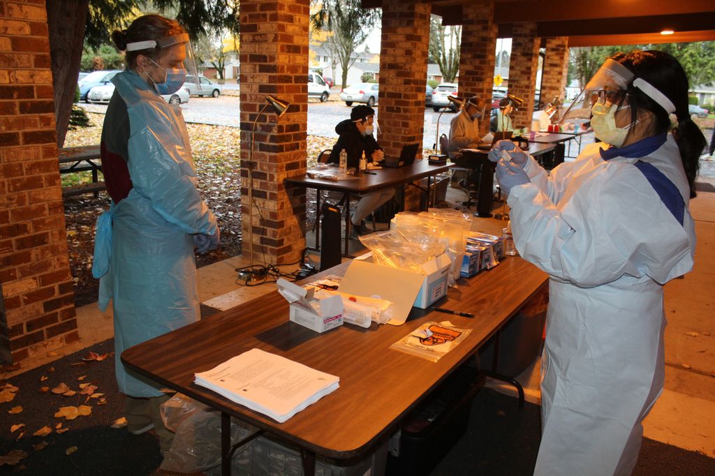 Two people clad in visors, masks and blue surgical gowns prepare testing materials at a table.  
