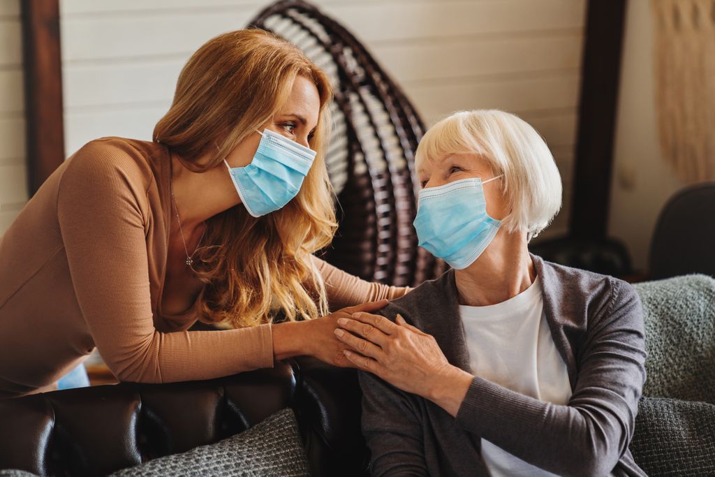a senior woman and her adult daughter wear protective face masks and clasp hands