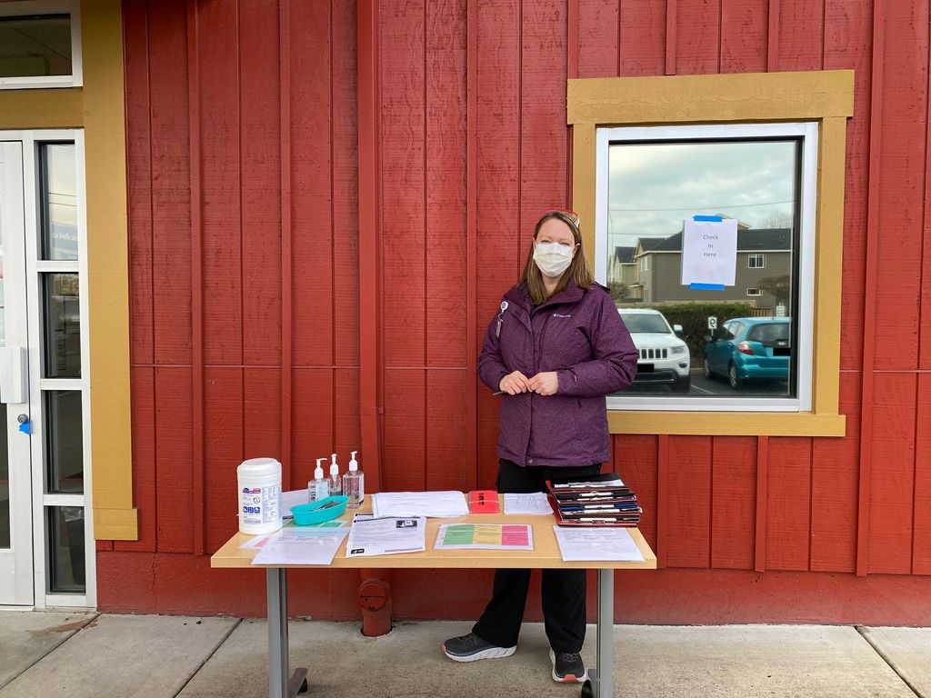 An adult woman in a protective mask stands at an information table in front of a red building; a sign on the window behind her reads 'check in here'