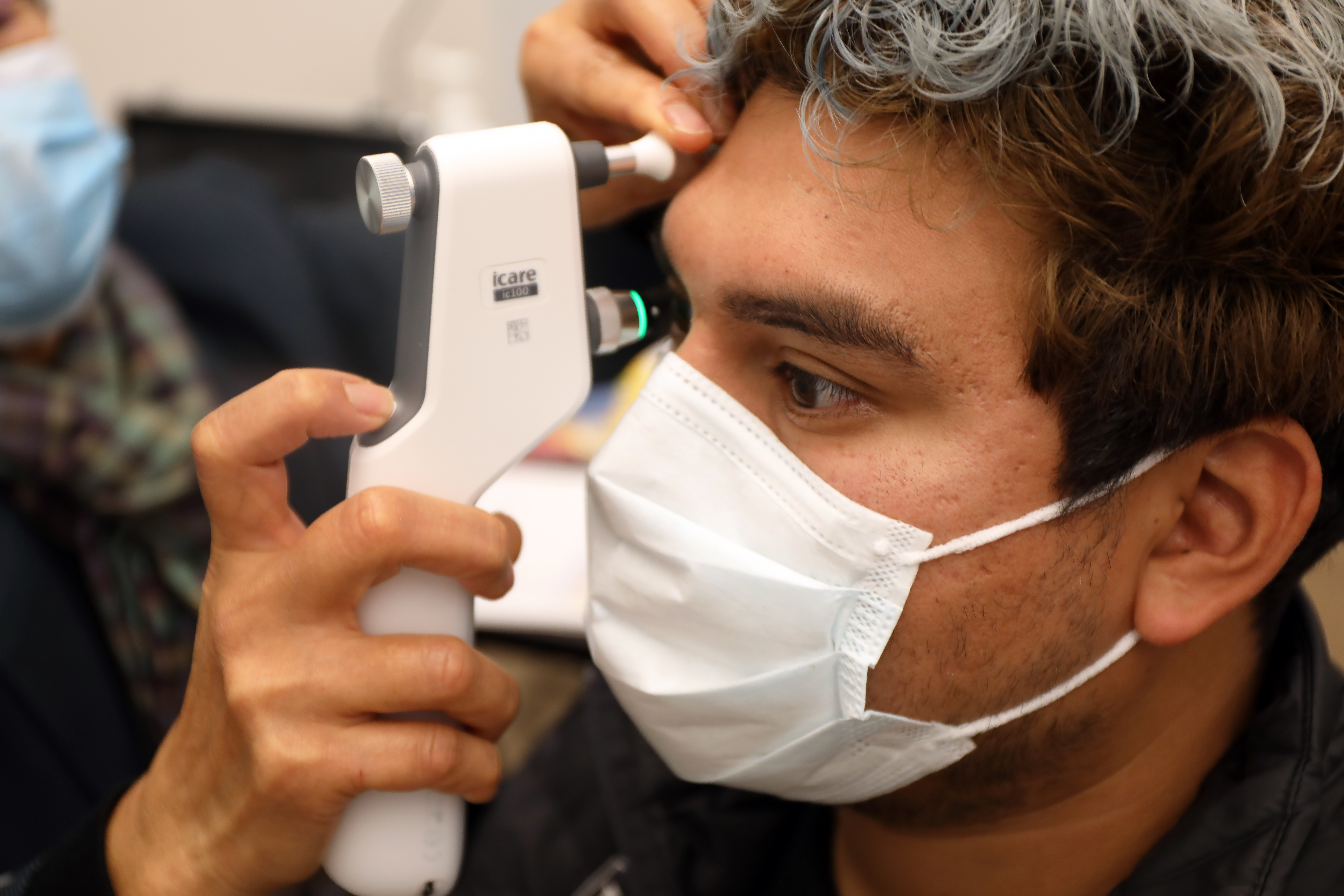A woman uses an eye pressure testing device close to the eye of Gerry Vasquez, who keeps his right eye open as the device is very close, he has dark hair and wearing a mask.
