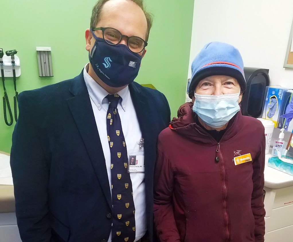 Flavio Rocha, M.D., a man with thin brown hair in a suit and mask, left, with patient Anne Matsen, a woman in a red jacket, blue beanie, and a face mask.