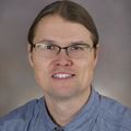 Erik Brodt, M.D. A person with long brown hair pulled back, wearing glasses and a blue shirt, smiling.