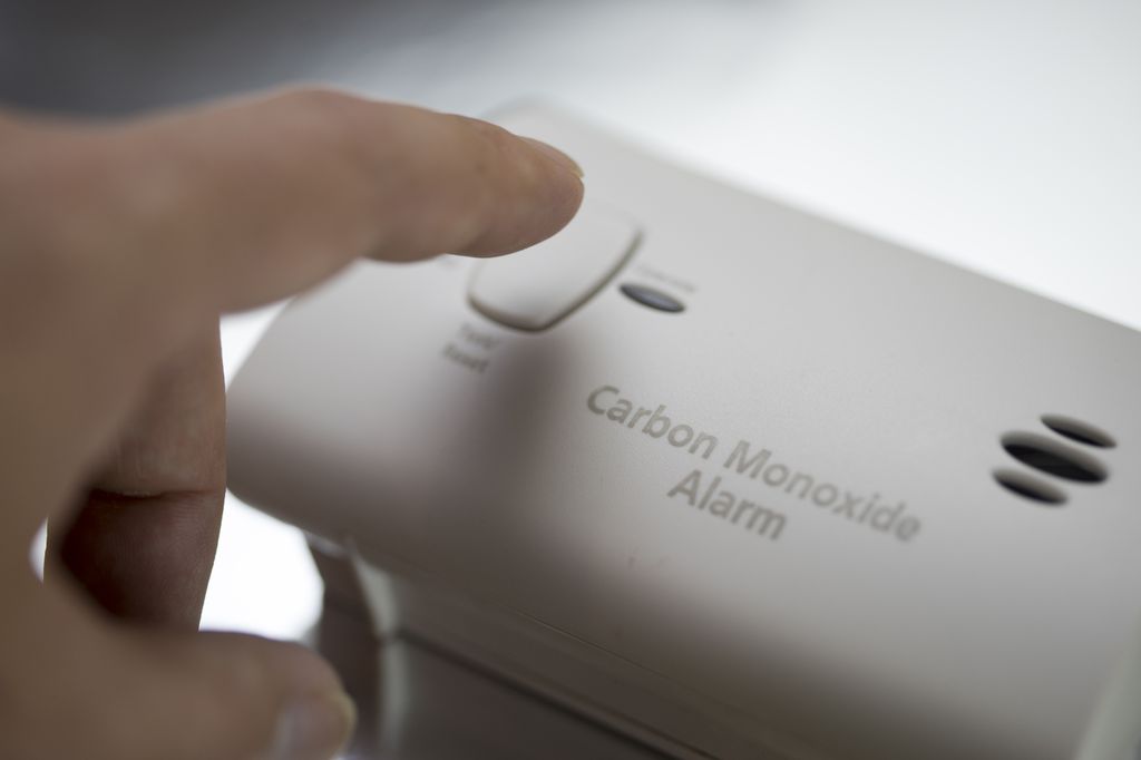 A carbon monoxide detector is a small battery-operated device that works similar to a smoke alarm. This image is of a person using their finder to press a button to test a carbon monoxide detector, a small white,  square device.