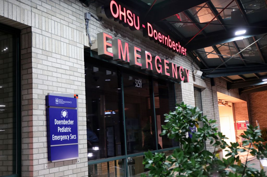 OHSU's emergency department exterior at night. Sign in red letters with a tinted window and a bench and bush outside.