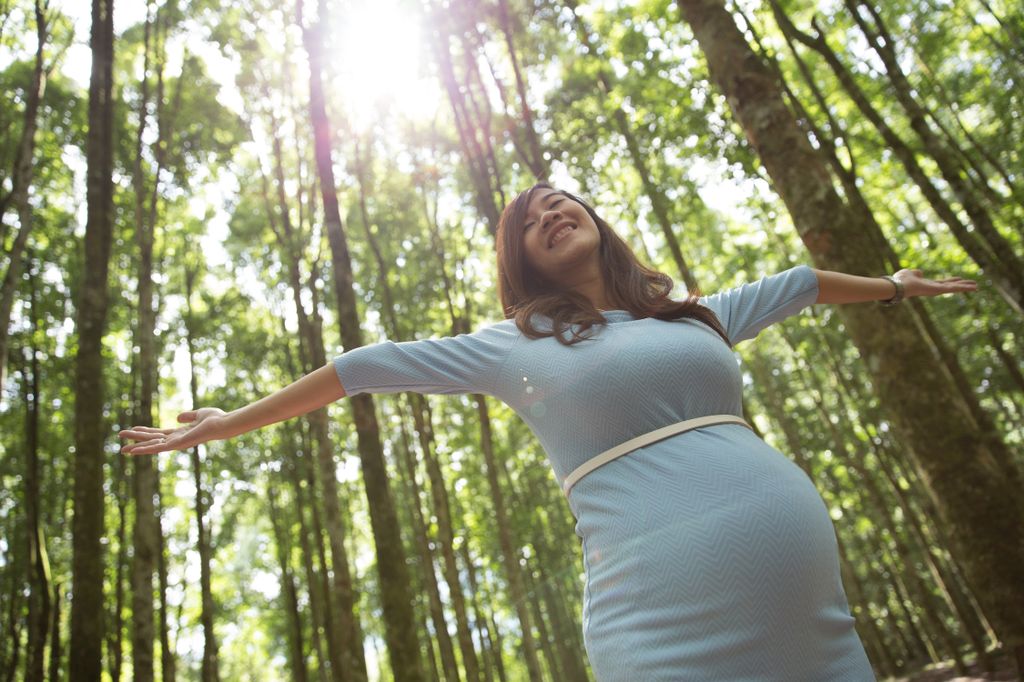 A pregnant woman with long dark hair stretches her arms open in a sunlit forest and smiles.