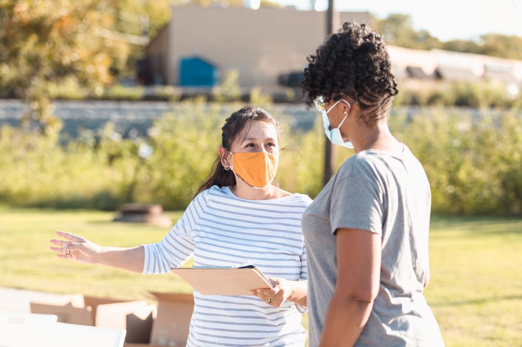 Two women with masks talk about health in their community, outdoors in a rural neighborhood.