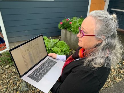Barb Snyder, a woman with gray, long hair pulled half-up and back, holding a laptop outside.
