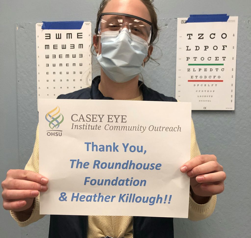 A woman wearing protective goggles and a face masks stands in front of two vision charts and holds a sign that reads "Thank you, The Roundhouse Foundation & Heather Killough!!"