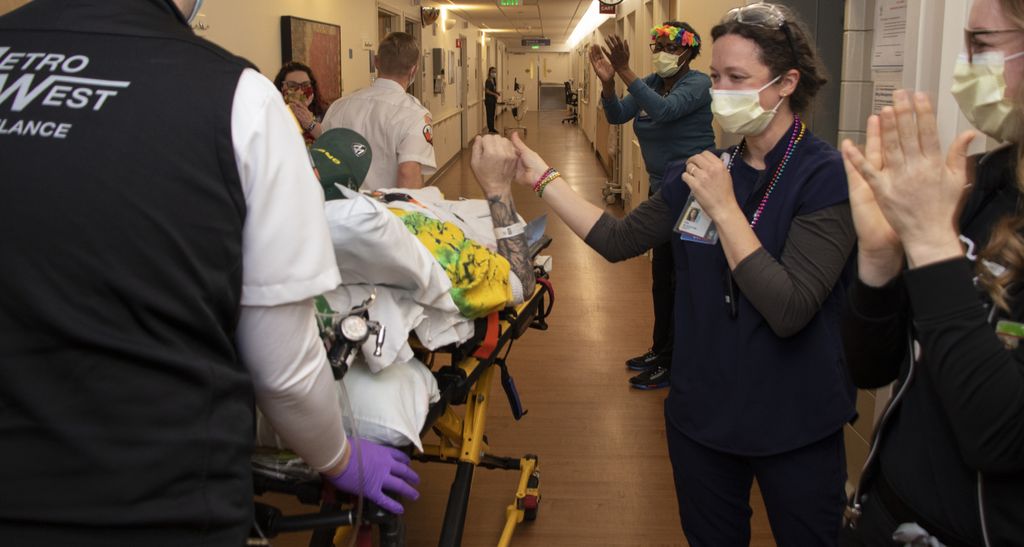 A health care worker wearing scrubs and Mardis Gras beads grasps the arm of a man laying in a stretcher who is being wheeled past a hallway lined with applauding staff. 