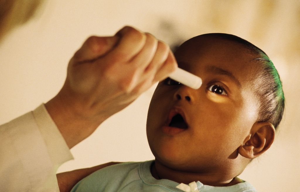 A brown-skinned infant receiving an eye exam, doctor's hand holds a light up and illuminates infant's left eye.