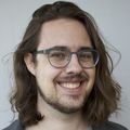 Headshot of Thomas Meath, a smiling young adult with long hair, glasses, and a beard