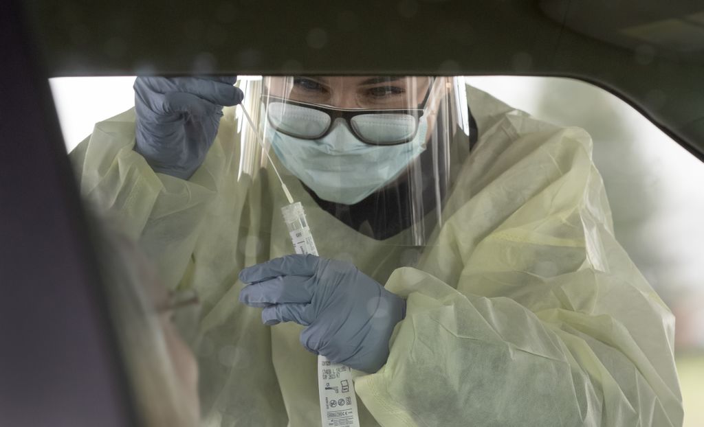 A woman leans into the car window as she peers through glasses and a clear face visor to insert a specimen swab into a plastic sleeve. 