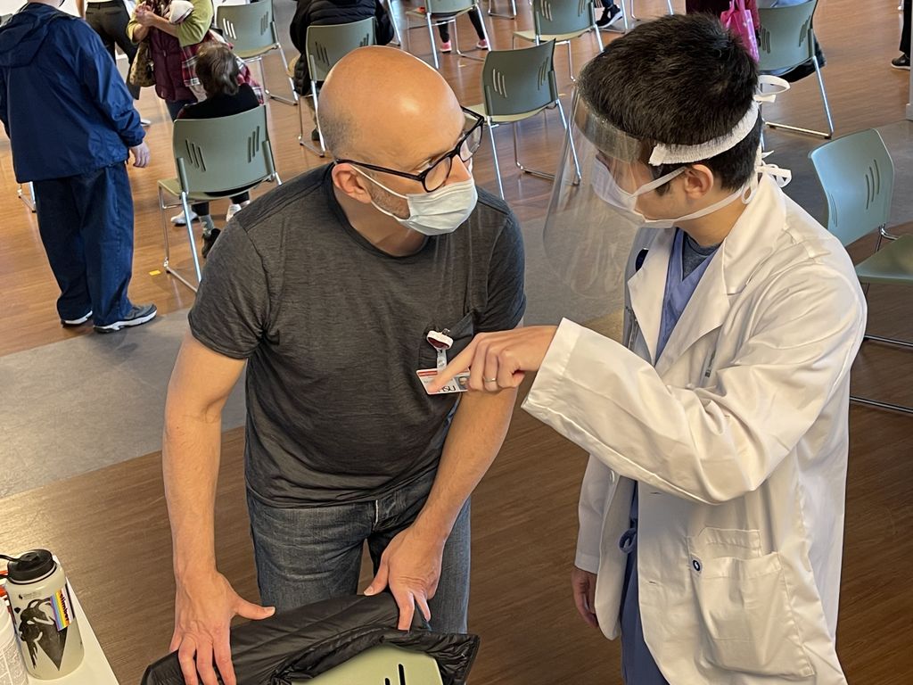 Two men stand talking: a bald white man with glasses and face mask (left), and an Asian with short black hair in a white lab coat and face shield.