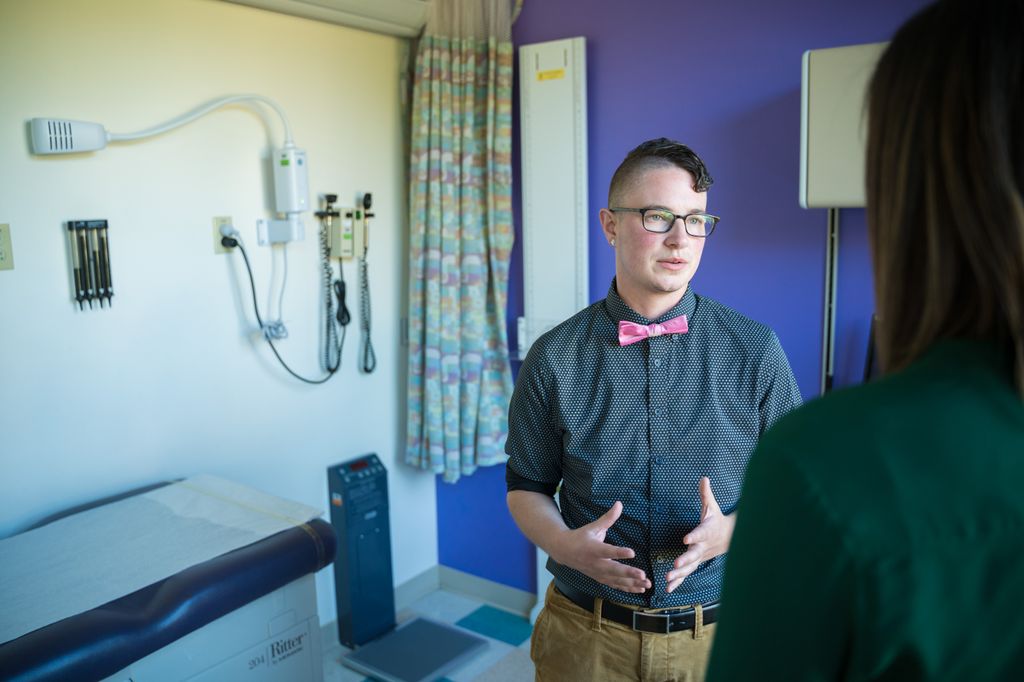 portrait of Jess Guerriero, M.A., M.S.W., an adult with glasses and a pink bow tie, standing in a medical office speaking to another adult (face not shown)