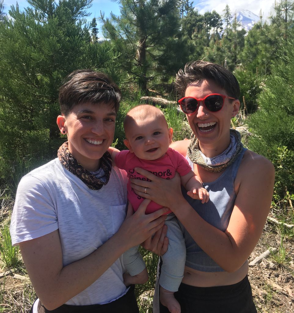Jae Downing and their wife hold their infant son, with evergreen trees in the background