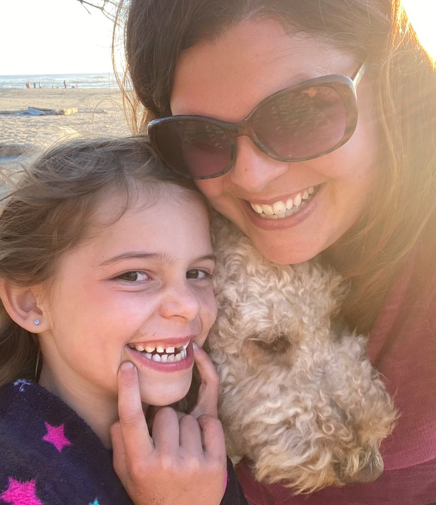 Brooke Hambly, a smiling adult with long brown hair wearing sunglasses, poses with her daughter - whose smile shows a missing front tooth - and their white, fuzzy dog