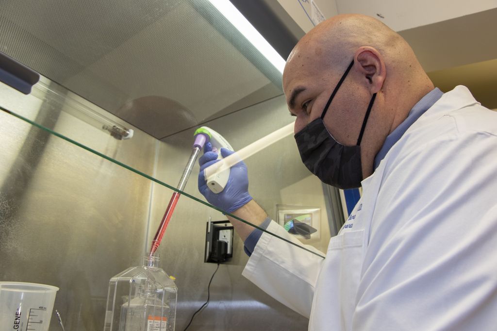 Ramon Barajas Jr., M.D., a bald, bearded man in a white lab coat, holding a large dropper filled with liquid above a container 