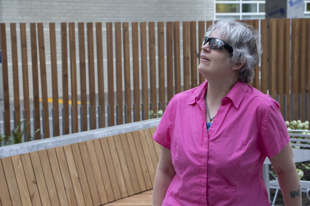 A woman with short, shaggy gray hear wears a bright pink, short-sleeved shirt and a pair of sunglasses as she looks up to the left.