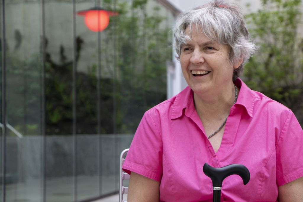 A woman with short, shaggy gray hear wears a bright pink, short-sleeved shirt as she smiles and looks toward the left.