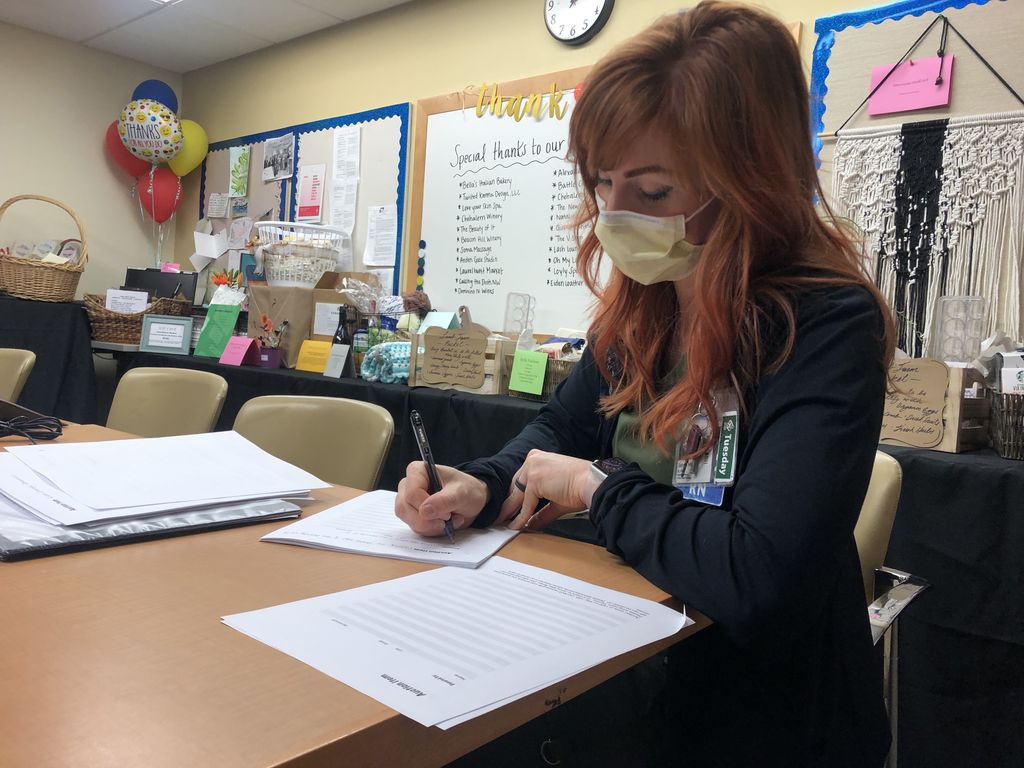 A woman with long red hair writes on pieces of paper, while tables full of goods up for auction sit in the background.