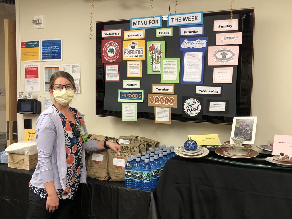 A woman wearing a face mask and colorful scrubs smiles as she picks up a brown-paper bag from a table with a row of similar bags.
