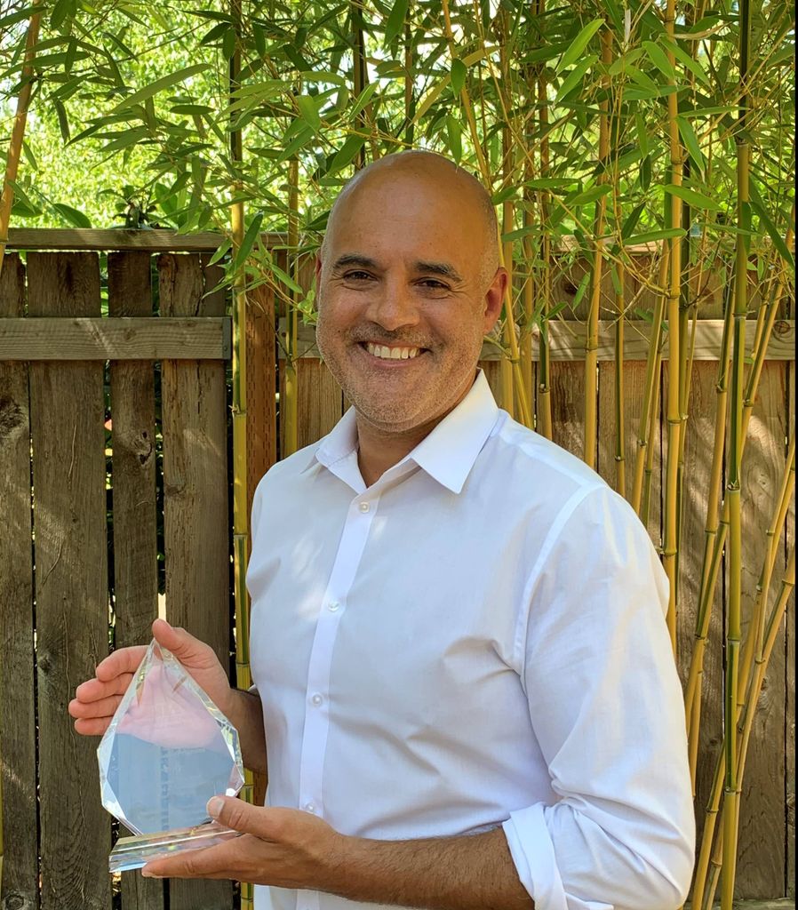 Cliff Coleman, M.D., M.P.H., a smiling bald man, stands in front of a fence and some tall bamboo, holding a crystal award