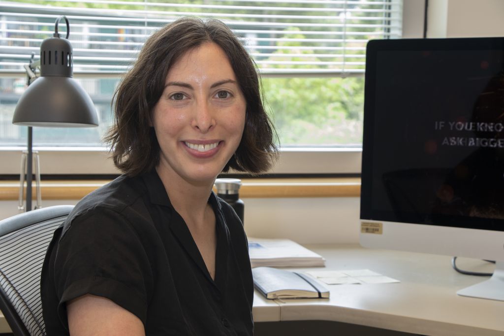 A woman with a short bob of dark hair and brown eyes sits in front of a computer and smiles at camera.