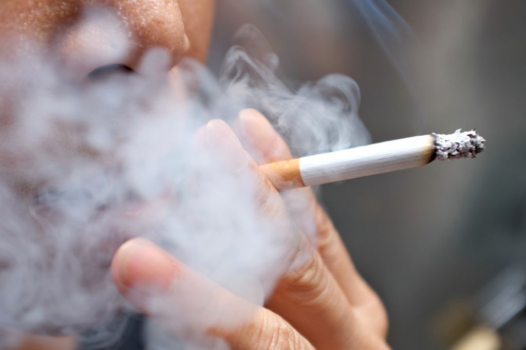 Close up of a person smoking a cigarette (only nose, mouth and hand visible) with smoke around them and a blurred background