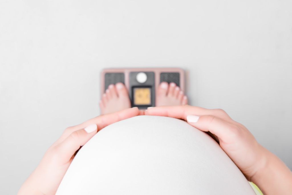 Pregnant woman with barefoot standing on weight scales. Hands touching big belly. Close up. Point of view shot. Top view.