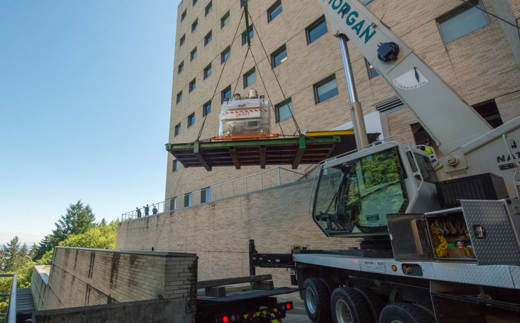 A crane outside of a large medical building hoists a massive magnet through the air.
