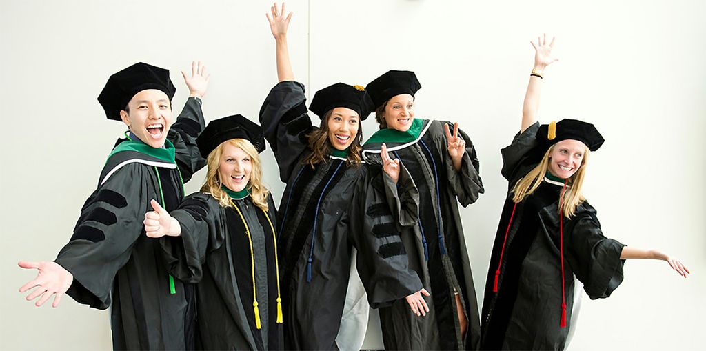 five young adult in graduation caps and gowns throw their hands in the air in celebration together, in front of a white background