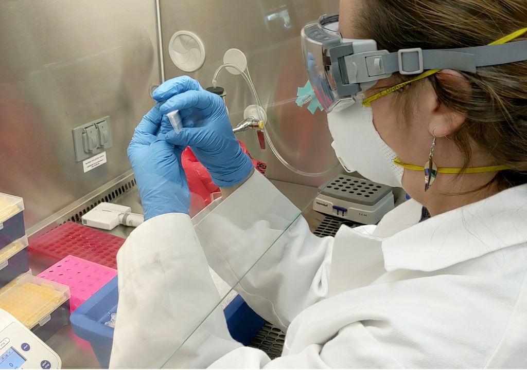 A female scientist in protective goggles and respirator mask examines samples in a row of small tubes, with lab equipment around.