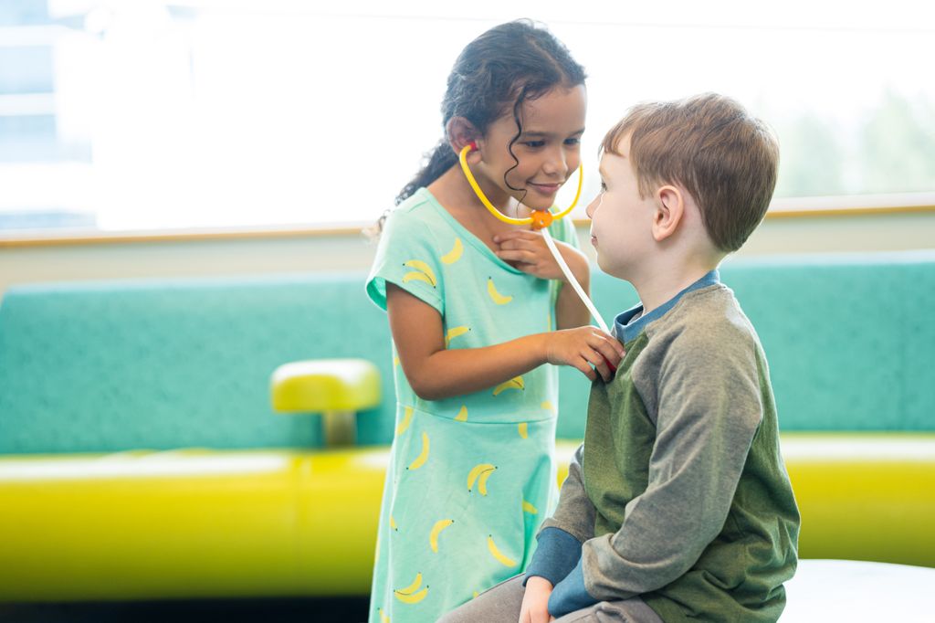 Two children play: a dark-haired girl wearing a toy stethoscope listens to a white boy's heart