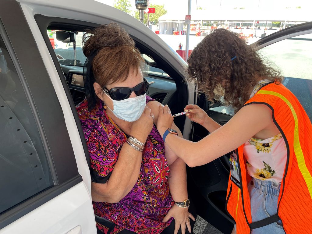 A woman seated on her car's seat exposes her upper arm and receives a vaccine injection from a curly-haired clinician in an orange safety vest.