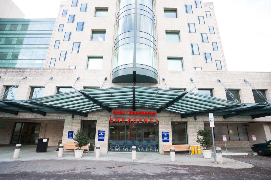 Architectural photo of OHSU's Emergency Department drop-off at the front of the hospital building. Building is made of pale stone, with a large glass awning and an exposed glass elevator shaft is above it.