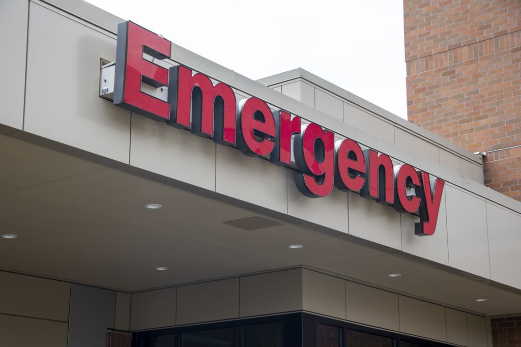 A sign with bold red letters reading “emergency” sits on the side of a beige and brick building.