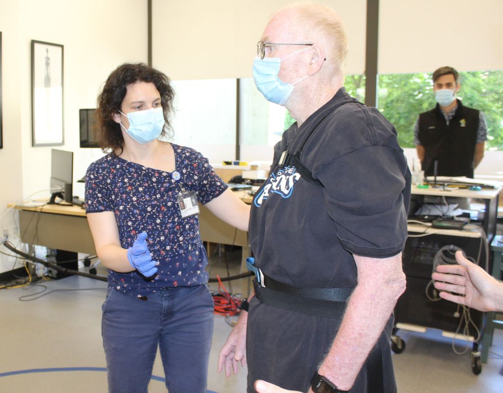 Marian Dale, M.D., M.C.R., a brunette woman in a medical face mask works with a senior male patient wearing straps across his chest, over his shoulders, and around his waist in a clinic setting.