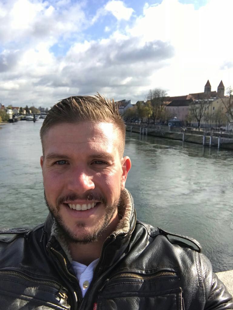 A bearded young man smiles while standing in front of a river.