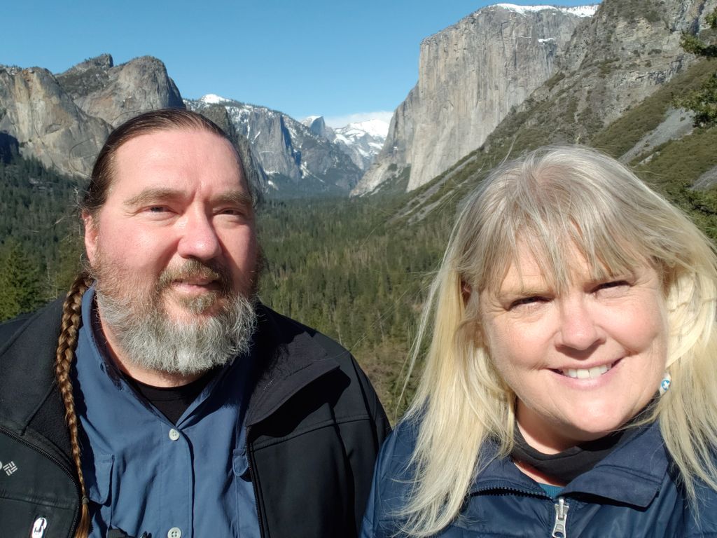 A bearded man and a blonde woman smile as they stand in front of snow-capped mountains and a green forest.
