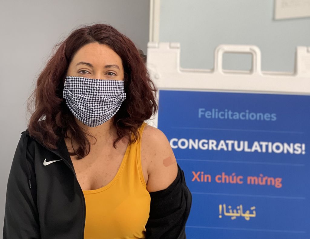 Young woman who just got vaccinated shows the Bandaid on her upper arm in front of a multilingual sign congratulating patients. 