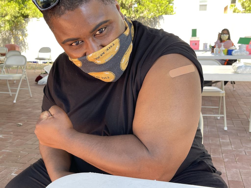 Forrest Taylor, a young Black man wearing a mask, flexes his left bicep while showing the bandage on his shoulder at the COVID vaccine injection site.