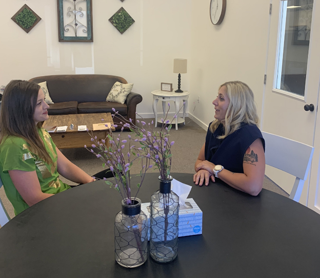 A young woman with long, brown hair sits talking with a smiling adult woman with blonde hair; they are seated at a black table with purple foliage in vases, with a couch in the background.
