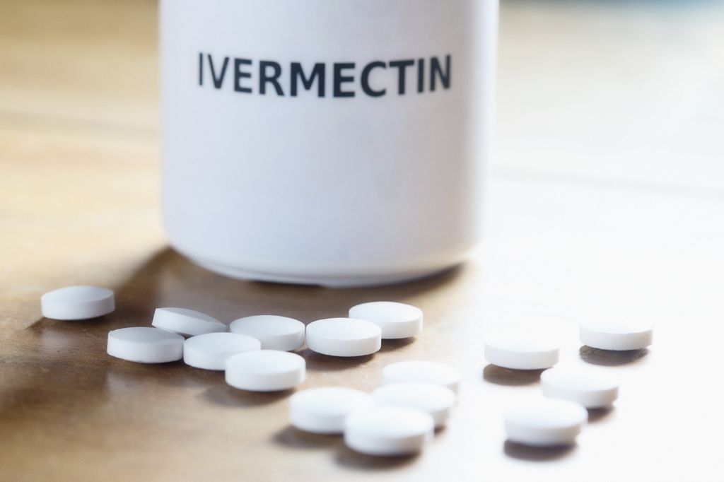 a white bottle labeled 'Ivermectin' sits on a wooden surface with about 15 unmarked white pills in front.