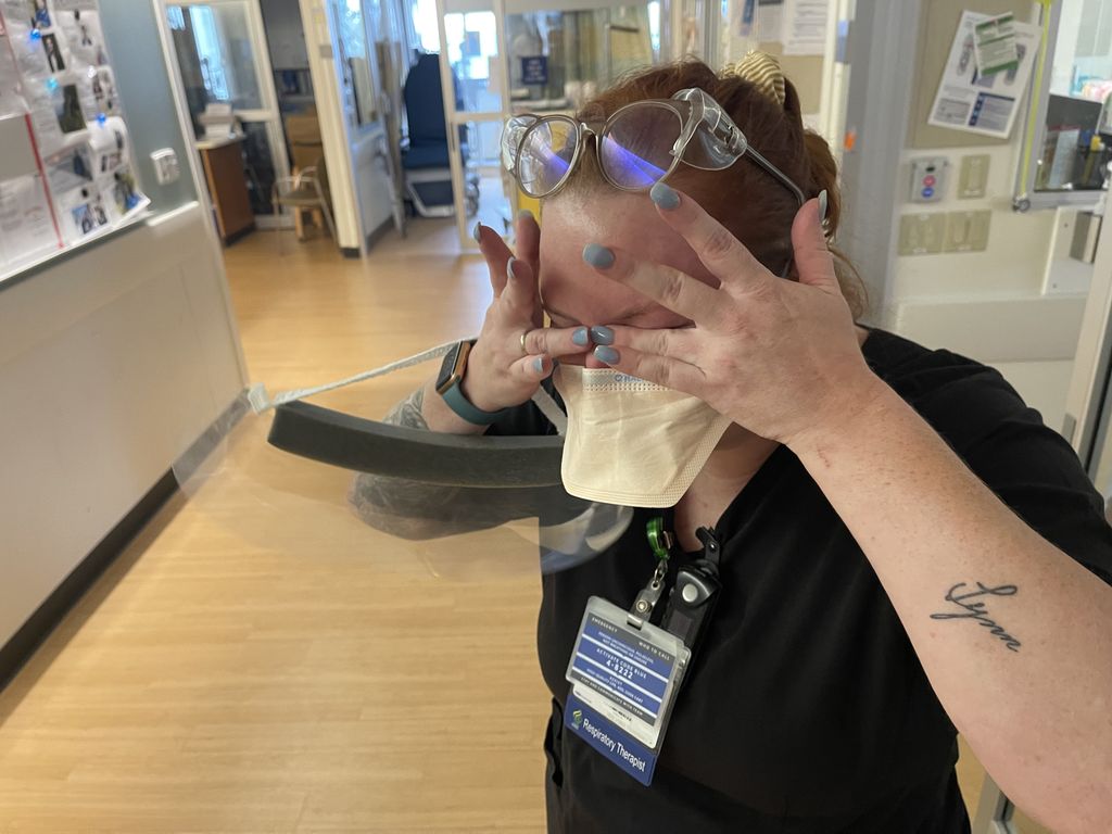 A crying nurse wipes their eyes around protective eyewear, and a face mask; she has a clear plastic face shield hanging from her arm.