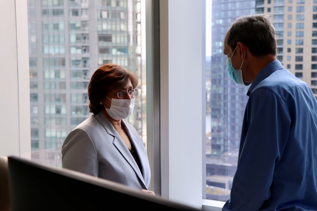 A tall man seen in profile speaks to a shorter woman; both wear surgical masks.