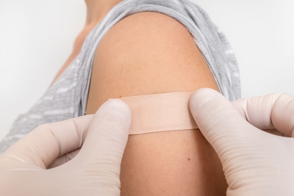  adhesive bandage being applied to a woman's left shoulder (shot from neck down, face not visible) by a pair of gloved hands