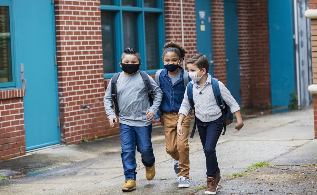 A group of three multi-ethnic elementary school students walking outside the school building, carrying backpacks, and wearing face masks. They are back to school during the covid-19 pandemic. The boy on the left is Hispanic. The one in the middle with long  hair tied back is mixed race African-American and Caucasian. And their friend on the right is mixed race Middle Eastern and Caucasian. They are 7, 10 and 8 years old.