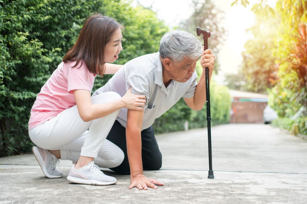 An elderly person falls on the sidewalk and another person is attempting to help them up.